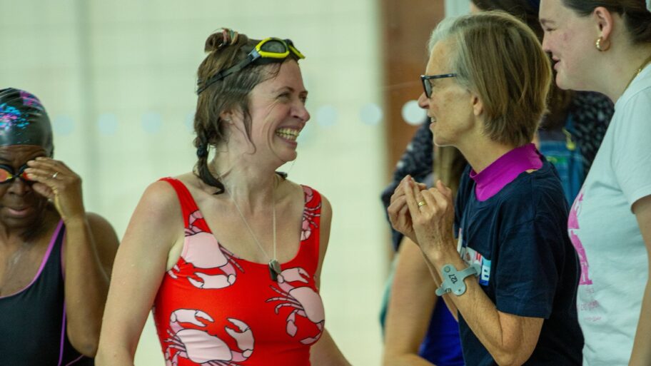 A female swimmer in a red costume laughing with two other women. She is wet and looks like she has just completed a race in the gala.